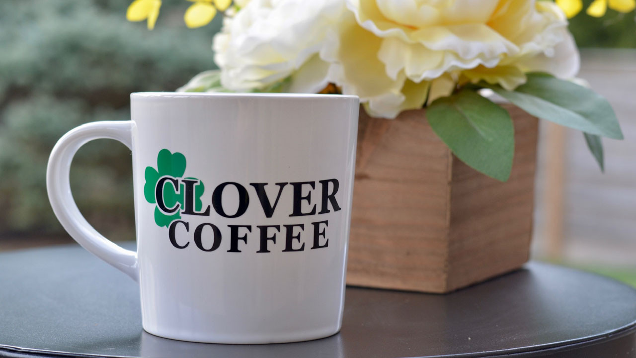 White mug with the text 'CLOVER COFFEE' and a green clover design, placed next to a wooden vase holding a white flower with a blurred green background.